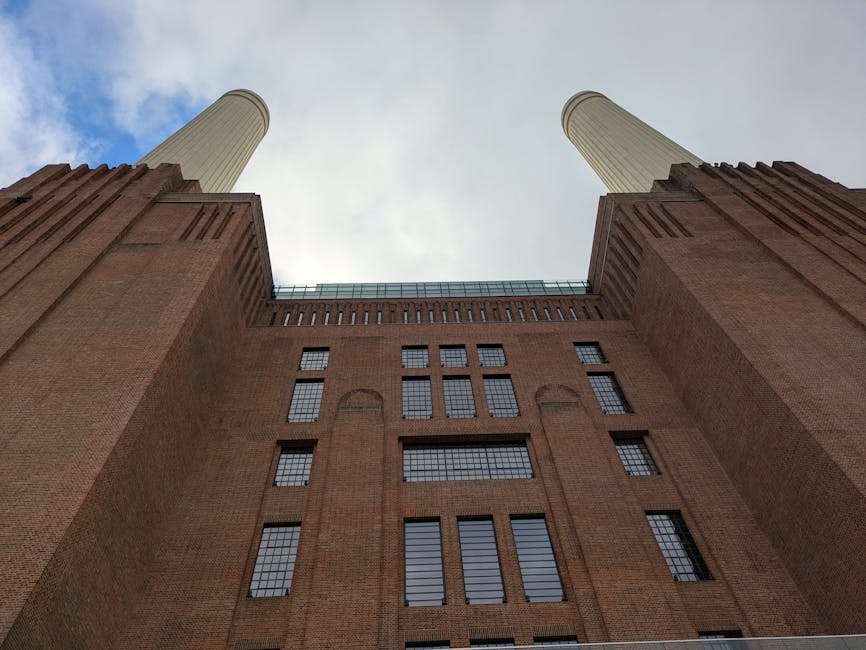View of the iconic Battersea Power Station, featuring a large brick facade with vertical narrow windows and a tall, white chimney extending into a clear blue sky. The exterior surface of the building 