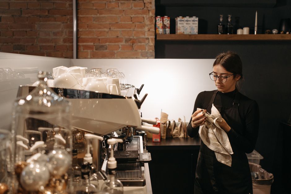 A person wearing glasses and a black shirt is performing surface cleaning in a modern lounge area, using a broom to sweep debris from the light wooden floor. The individual is dressed with an apron, i